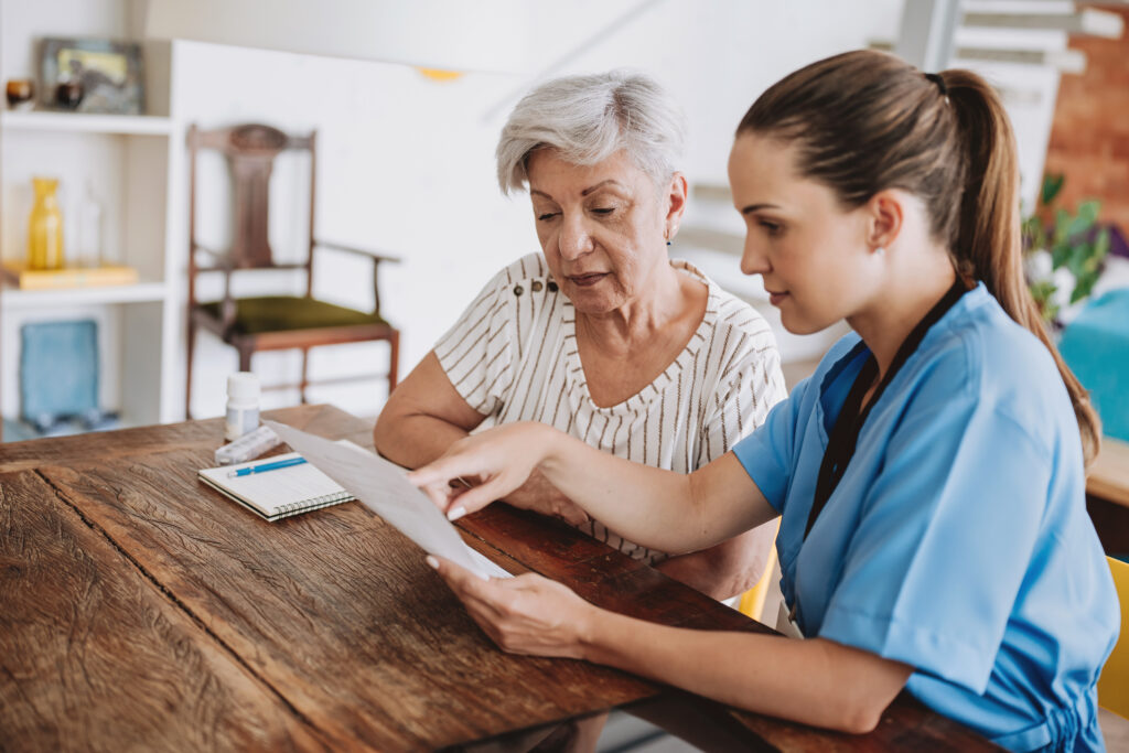 A caregiver sits with a senior woman at a wooden table, reviewing documents together in a bright home setting — representing planning, independence, and confidence in ageing in place.