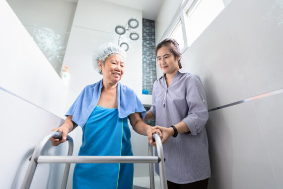 A caregiver gently supports an elderly woman using a walker in a bright bathroom, ensuring safety and comfort during her bath — illustrating compassionate Alzheimer’s care.