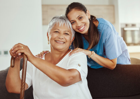 A smiling caregiver gently rests her hands on an older woman’s shoulders, both sharing a warm connection that reflects compassion, trust, and quality in-home care.