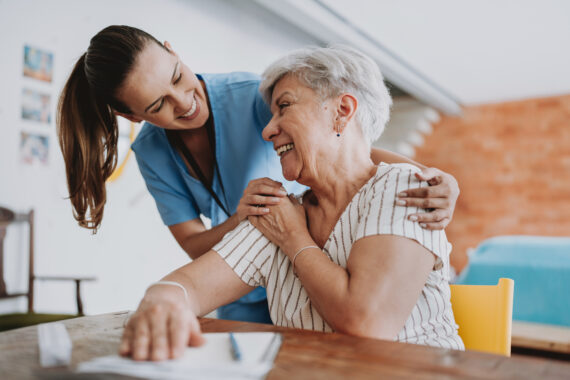 Smiling elderly woman sitting at her kitchen table while a caregiver gently supports her with a hand on her arm and shoulder in a warm, welcoming home environment.