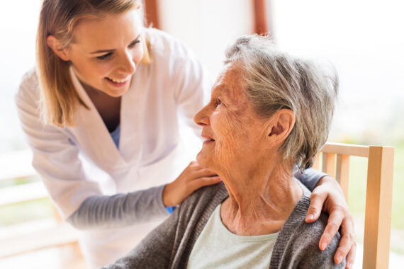 A smiling caregiver gently placing a hand on an older woman’s shoulder while they share a warm, supportive moment at home.
